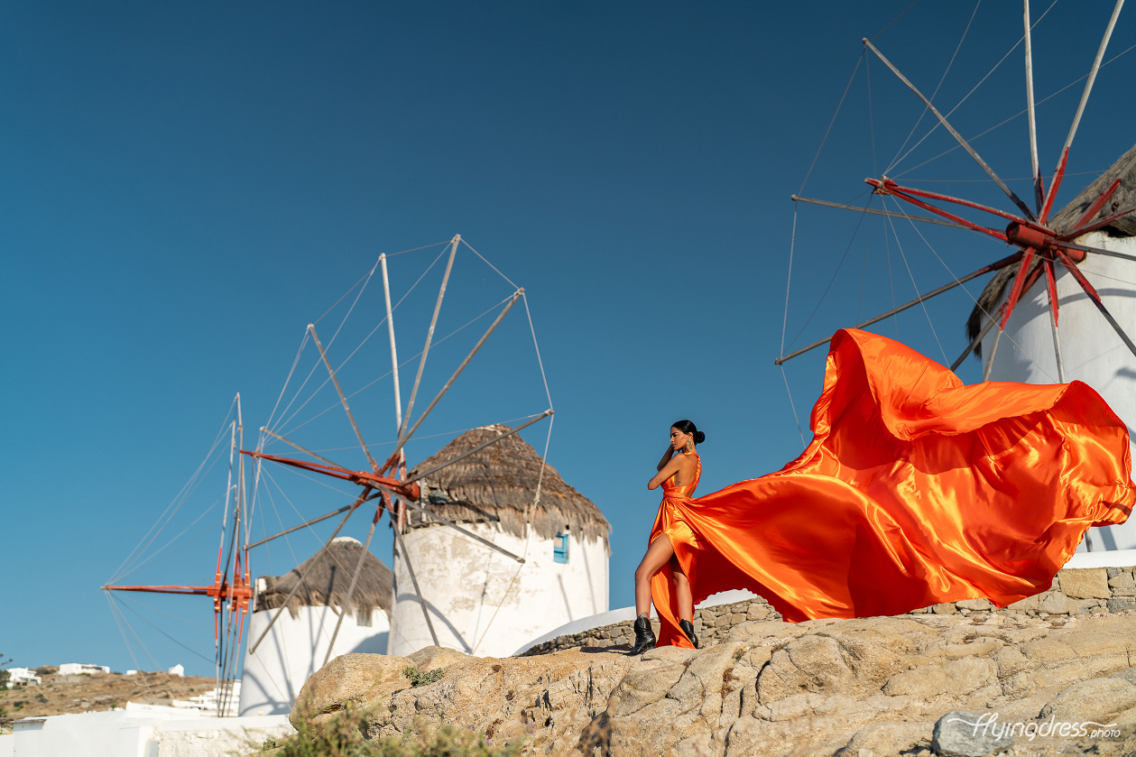 A woman in a vibrant orange dress poses on a rocky hill with the traditional windmills of Mykonos in the background, her dress billowing dramatically in the wind against a clear blue sky.